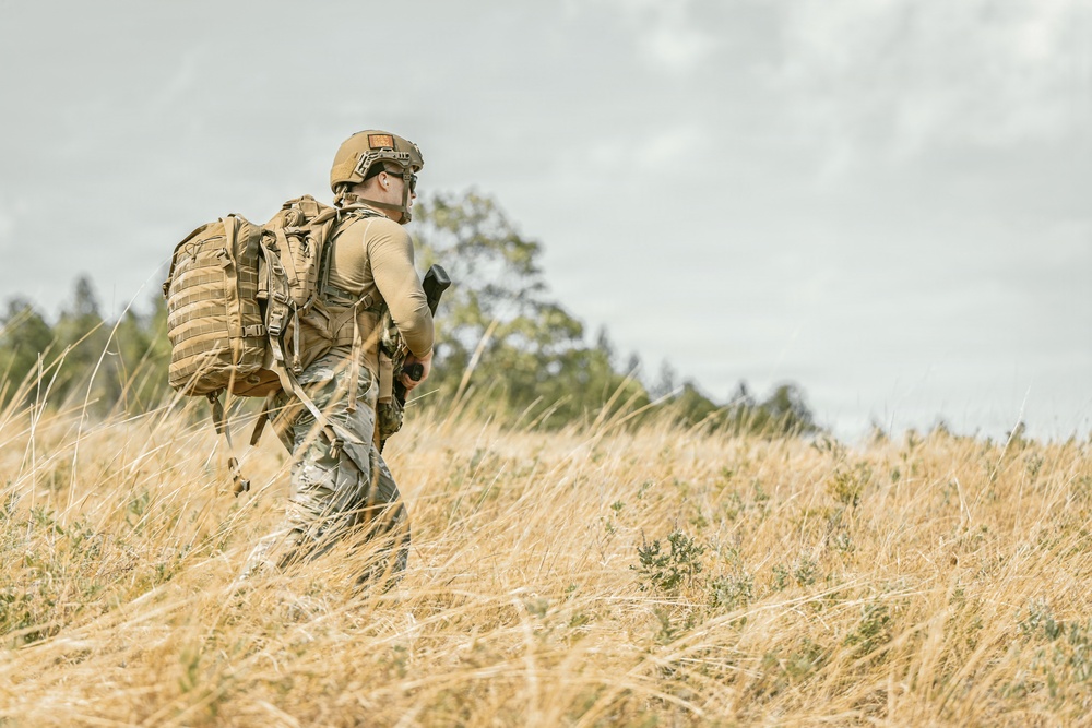 A soldier or tactical operator is walking through a dry, golden grassland field. The individual is equipped with full tactical gear including a helmet, body armor, and a large MOLLE-style backpack. The scene appears to be an outdoor field exercise or patrol in a temperate environment.