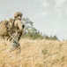 A soldier or tactical operator is walking through a dry, golden grassland field. The individual is equipped with full tactical gear including a helmet, body armor, and a large MOLLE-style backpack. The scene appears to be an outdoor field exercise or patrol in a temperate environment.