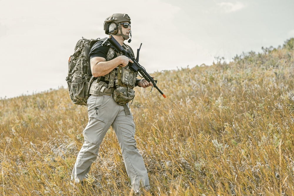 A heavily equipped individual is walking through an open field of dry golden grass. The person is wearing full tactical gear including a helmet, body armor vest, large backpack, and carrying a rifle. The setting appears to be an outdoor training or operational environment under an overcast sky.