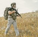 A heavily equipped individual is walking through an open field of dry golden grass. The person is wearing full tactical gear including a helmet, body armor vest, large backpack, and carrying a rifle. The setting appears to be an outdoor training or operational environment under an overcast sky.