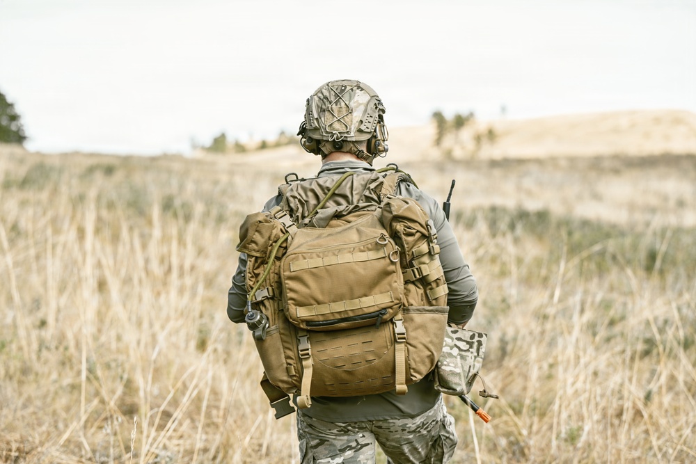 A soldier or tactical operator is seen from behind, standing in an open dry grassland field. The individual is fully equipped with military-grade gear including a tactical backpack, helmet, and communications equipment. The scene appears to be a field exercise or patrol in a rural, arid environment.