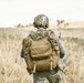 A soldier or tactical operator is seen from behind, standing in an open dry grassland field. The individual is fully equipped with military-grade gear including a tactical backpack, helmet, and communications equipment. The scene appears to be a field exercise or patrol in a rural, arid environment.