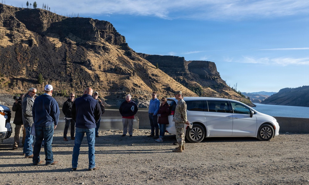 A group of approximately 10-12 people are gathered in a gravel parking or overlook area near a large river or reservoir, with dramatic basalt cliffs and canyon walls in the background. One individual is wearing military camouflage uniform, suggesting this may be an official site visit or briefing. A white minivan (appears to be a Chrysler Pacifica) is parked nearby, and one person is holding a camera.