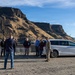 A group of approximately 10-12 people are gathered in a gravel parking or overlook area near a large river or reservoir, with dramatic basalt cliffs and canyon walls in the background. One individual is wearing military camouflage uniform, suggesting this may be an official site visit or briefing. A white minivan (appears to be a Chrysler Pacifica) is parked nearby, and one person is holding a camera.