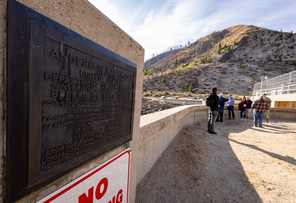 A bronze commemorative plaque for Anderson Ranch Dam, built by the United States Department of the Interior Bureau of Reclamation, is mounted on a concrete structure in the foreground. The dam's specifications are visible on the plaque, including a height of 456 feet, length of 1350 feet, and storage capacity of 500,000 acre feet. In the background, a group of people stand on what appears to be the dam structure, with a rocky, partially burned mountainside visible behind them.