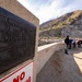 A bronze commemorative plaque for Anderson Ranch Dam, built by the United States Department of the Interior Bureau of Reclamation, is mounted on a concrete structure in the foreground. The dam's specifications are visible on the plaque, including a height of 456 feet, length of 1350 feet, and storage capacity of 500,000 acre feet. In the background, a group of people stand on what appears to be the dam structure, with a rocky, partially burned mountainside visible behind them.