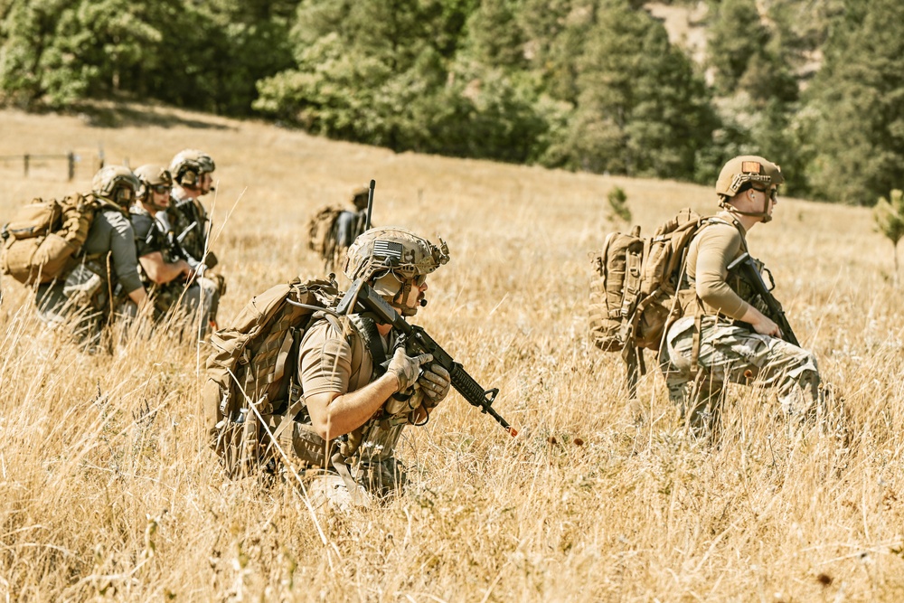 A group of military or tactical personnel are moving through a dry grassland field with a forested hillside in the background. The individuals are equipped with full tactical gear including helmets, body armor, large rucksacks, and firearms. The scene appears to depict a field training exercise or patrol movement.