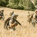A group of military or tactical personnel are moving through a dry grassland field with a forested hillside in the background. The individuals are equipped with full tactical gear including helmets, body armor, large rucksacks, and firearms. The scene appears to depict a field training exercise or patrol movement.