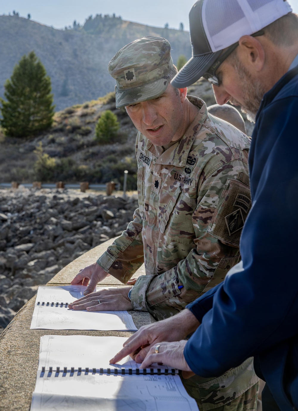 A U.S. Army officer wearing OCP (Operational Camouflage Pattern) uniform with the name tape 'DROESE' and a Lieutenant Colonel oak leaf insignia reviews spiral-bound documents or blueprints with a civilian man outdoors. They appear to be conducting a site inspection or project review at what looks like a dam or water infrastructure facility. The background shows rocky terrain, sparse vegetation, and mountainous landscape.