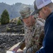 A U.S. Army officer wearing OCP (Operational Camouflage Pattern) uniform with the name tape 'DROESE' and a Lieutenant Colonel oak leaf insignia reviews spiral-bound documents or blueprints with a civilian man outdoors. They appear to be conducting a site inspection or project review at what looks like a dam or water infrastructure facility. The background shows rocky terrain, sparse vegetation, and mountainous landscape.