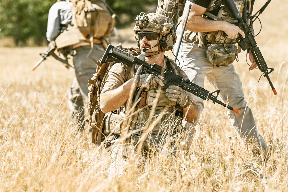 Three individuals in military-style tactical gear are positioned in a dry grassfield. The central figure is kneeling and holding an assault rifle with the name 'GULLAGE' visible on the stock, wearing a helmet with communications equipment. The scene appears to be a training exercise or tactical drill based on the orange safety tip visible on one of the weapons.