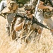 Three individuals in military-style tactical gear are positioned in a dry grassfield. The central figure is kneeling and holding an assault rifle with the name 'GULLAGE' visible on the stock, wearing a helmet with communications equipment. The scene appears to be a training exercise or tactical drill based on the orange safety tip visible on one of the weapons.