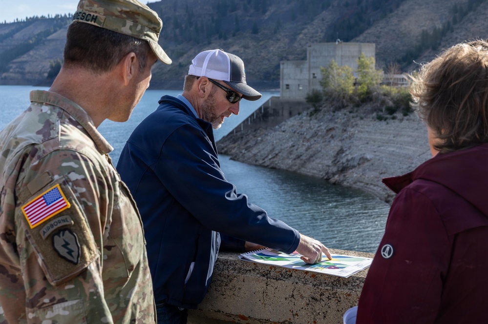 Three individuals are gathered on what appears to be a dam structure, reviewing printed documents or plans. One person is in U.S. Army military uniform with Airborne and unit patches visible, while the other two are in civilian attire. The setting includes a reservoir or lake with a concrete dam infrastructure and mountainous terrain in the background.