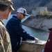 Three individuals are gathered on what appears to be a dam structure, reviewing printed documents or plans. One person is in U.S. Army military uniform with Airborne and unit patches visible, while the other two are in civilian attire. The setting includes a reservoir or lake with a concrete dam infrastructure and mountainous terrain in the background.