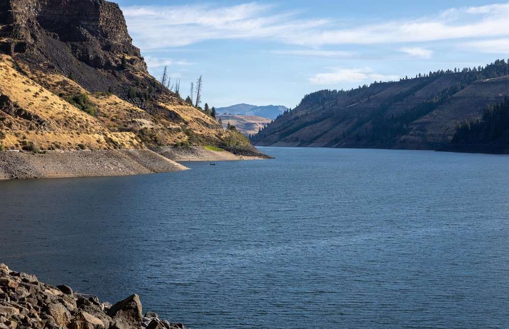 A wide river or reservoir winds through a steep, arid canyon landscape with dry golden-brown hillsides and rocky cliffs. The water is calm and deep blue, flanked by exposed rocky shorelines suggesting a below-normal water level. Sparse conifer trees and dead snags dot the hillsides under a partly cloudy sky.