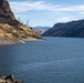 A wide river or reservoir winds through a steep, arid canyon landscape with dry golden-brown hillsides and rocky cliffs. The water is calm and deep blue, flanked by exposed rocky shorelines suggesting a below-normal water level. Sparse conifer trees and dead snags dot the hillsides under a partly cloudy sky.