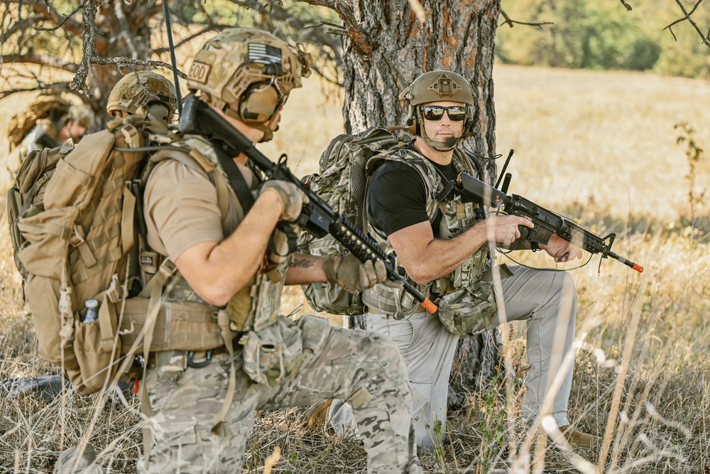 Two armed individuals in tactical military-style gear are positioned near a tree in an open field or woodland environment. Both are equipped with rifles, helmets, backpacks, and communication headsets, suggesting a training exercise or tactical operation. A third individual is partially visible in the background.