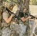 Two armed individuals in tactical military-style gear are positioned near a tree in an open field or woodland environment. Both are equipped with rifles, helmets, backpacks, and communication headsets, suggesting a training exercise or tactical operation. A third individual is partially visible in the background.