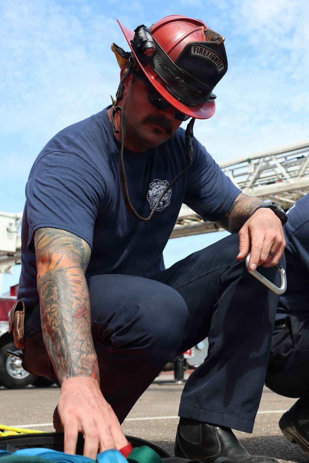 A firefighter wearing a red helmet labeled 'FIREFIGHTER' is crouched down and handling equipment, including a carabiner clip. He is dressed in a navy blue t-shirt with a 'Naval Base Coronado' logo and navy work pants. His left arm features extensive colorful tattoos, and he appears to be preparing or checking gear outdoors.