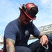 A firefighter wearing a red helmet labeled 'FIREFIGHTER' is crouched down and handling equipment, including a carabiner clip. He is dressed in a navy blue t-shirt with a 'Naval Base Coronado' logo and navy work pants. His left arm features extensive colorful tattoos, and he appears to be preparing or checking gear outdoors.
