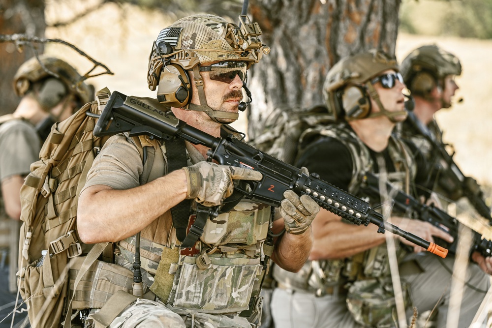The image shows multiple individuals in full tactical military-style gear, including camouflage uniforms, plate carriers, helmets with accessories, and communications headsets. They are holding assault-style rifles and appear to be in a staged or training exercise environment near trees in an arid outdoor setting. The foreground subject has a name tag reading 'Gullage' and an American flag patch visible on their helmet.