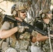 The image shows multiple individuals in full tactical military-style gear, including camouflage uniforms, plate carriers, helmets with accessories, and communications headsets. They are holding assault-style rifles and appear to be in a staged or training exercise environment near trees in an arid outdoor setting. The foreground subject has a name tag reading 'Gullage' and an American flag patch visible on their helmet.