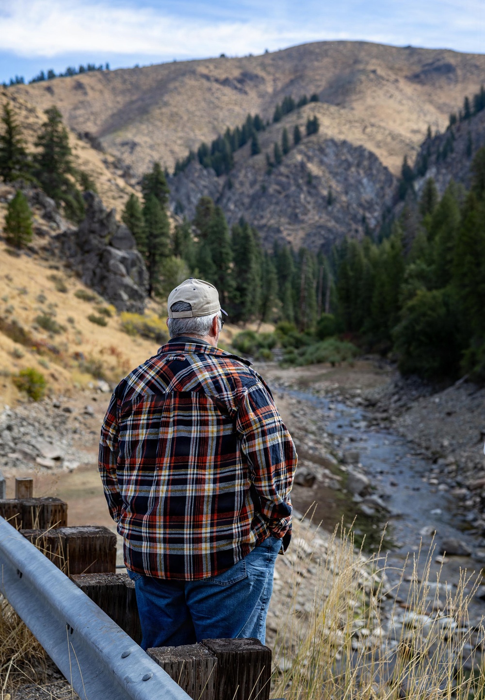 An older man wearing a plaid flannel shirt, jeans, and a baseball cap stands with his back to the camera, looking out over a shallow rocky creek winding through a mountain valley. The landscape features dry golden hillsides, conifer trees, and rugged rocky terrain under a partly cloudy sky. He appears to be standing on or near a small bridge with a metal guardrail and wooden posts.