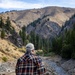 An older man wearing a plaid flannel shirt, jeans, and a baseball cap stands with his back to the camera, looking out over a shallow rocky creek winding through a mountain valley. The landscape features dry golden hillsides, conifer trees, and rugged rocky terrain under a partly cloudy sky. He appears to be standing on or near a small bridge with a metal guardrail and wooden posts.
