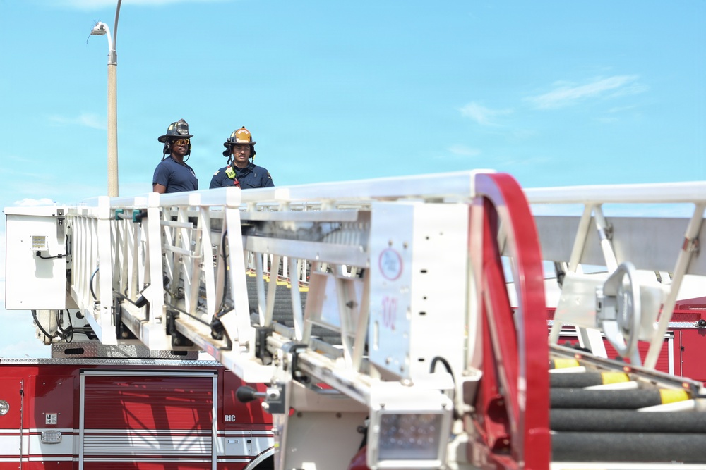 Two firefighters in full gear, including helmets and navy uniforms, are positioned on the aerial ladder platform of a red fire truck. The image appears to be taken during a training exercise or active deployment, with the extended ladder mechanism prominently visible. The scene is set against a clear blue sky with a street lamp visible in the background.