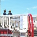 Two firefighters in full gear, including helmets and navy uniforms, are positioned on the aerial ladder platform of a red fire truck. The image appears to be taken during a training exercise or active deployment, with the extended ladder mechanism prominently visible. The scene is set against a clear blue sky with a street lamp visible in the background.