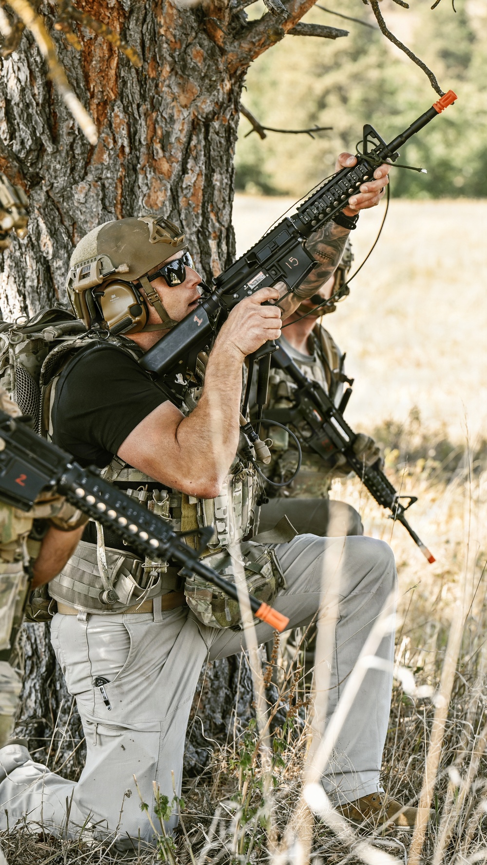 Two individuals in tactical gear are positioned behind a tree in an outdoor field environment, holding rifles with orange muzzle safety caps. Both are wearing camouflage and tactical equipment including plate carriers and pouches. The orange barrel blockers and numbered markers on the rifles suggest this is a training exercise or simulation event rather than live combat.