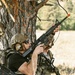 Two individuals in tactical gear are positioned behind a tree in an outdoor field environment, holding rifles with orange muzzle safety caps. Both are wearing camouflage and tactical equipment including plate carriers and pouches. The orange barrel blockers and numbered markers on the rifles suggest this is a training exercise or simulation event rather than live combat.