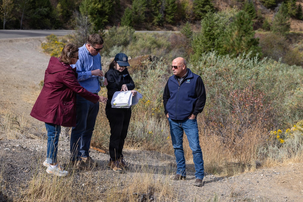Four adults are gathered outdoors in a dry, semi-arid landscape reviewing documents or plans. Three of the individuals are clustered together examining papers, while a fourth person stands nearby observing. The setting appears to be a field or site visit, possibly related to land assessment, environmental review, or project planning.