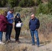 Four adults are gathered outdoors in a dry, semi-arid landscape reviewing documents or plans. Three of the individuals are clustered together examining papers, while a fourth person stands nearby observing. The setting appears to be a field or site visit, possibly related to land assessment, environmental review, or project planning.