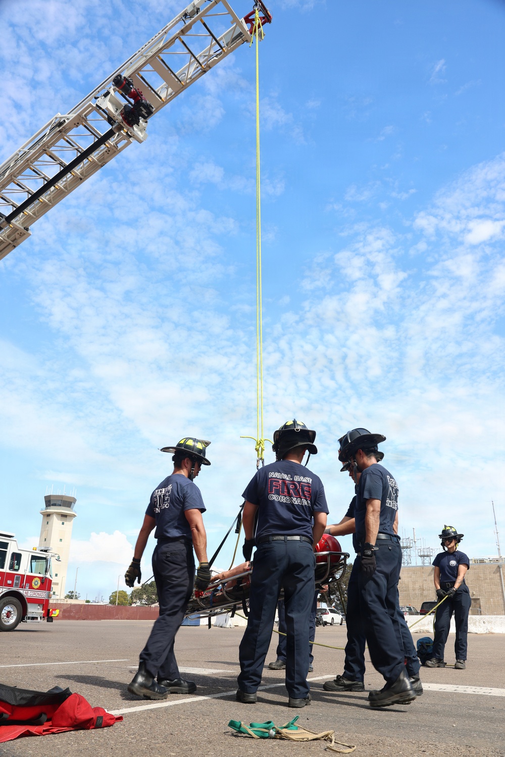 Firefighters from Naval Base Coronado are conducting a high-angle rescue training exercise using an extended aerial ladder truck. A team of approximately five personnel on the ground are managing a rescue stretcher/basket attached to a yellow rope running up to the top of the ladder. The exercise takes place on a paved area near what appears to be an airfield control tower.