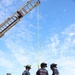 Firefighters from Naval Base Coronado are conducting a high-angle rescue training exercise using an extended aerial ladder truck. A team of approximately five personnel on the ground are managing a rescue stretcher/basket attached to a yellow rope running up to the top of the ladder. The exercise takes place on a paved area near what appears to be an airfield control tower.