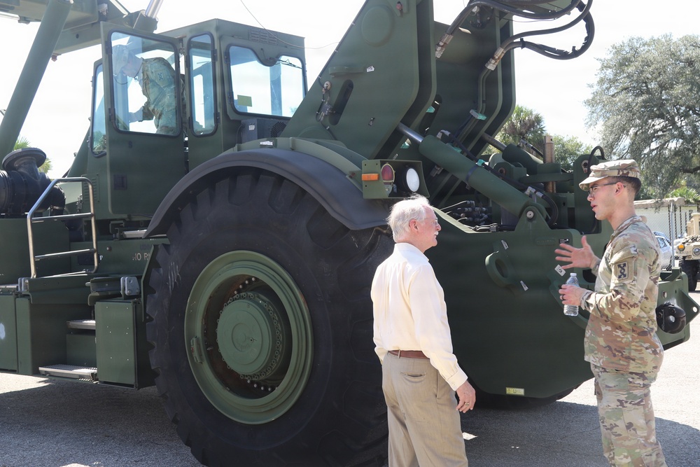 A U.S. Army soldier in OCP (Operational Camouflage Pattern) uniform is briefing a civilian man in front of a large military olive-drab green heavy equipment vehicle, likely a rough terrain forklift or container handler. The soldier appears to be explaining features of the vehicle while holding a water bottle. The setting appears to be an outdoor military installation or equipment demonstration area.