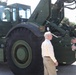 A U.S. Army soldier in OCP (Operational Camouflage Pattern) uniform is briefing a civilian man in front of a large military olive-drab green heavy equipment vehicle, likely a rough terrain forklift or container handler. The soldier appears to be explaining features of the vehicle while holding a water bottle. The setting appears to be an outdoor military installation or equipment demonstration area.