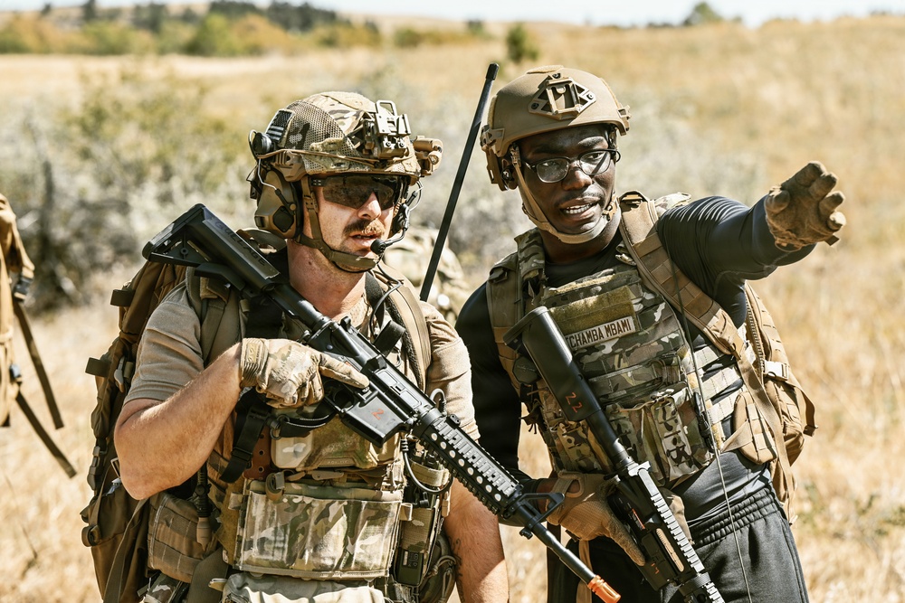 Two armed military personnel in tactical gear are standing in an open field with dry grassland in the background. One individual is pointing in a direction, appearing to communicate or coordinate with the other. Both are equipped with rifles, body armor, helmets, and tactical accessories.