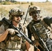 Two armed military personnel in tactical gear are standing in an open field with dry grassland in the background. One individual is pointing in a direction, appearing to communicate or coordinate with the other. Both are equipped with rifles, body armor, helmets, and tactical accessories.