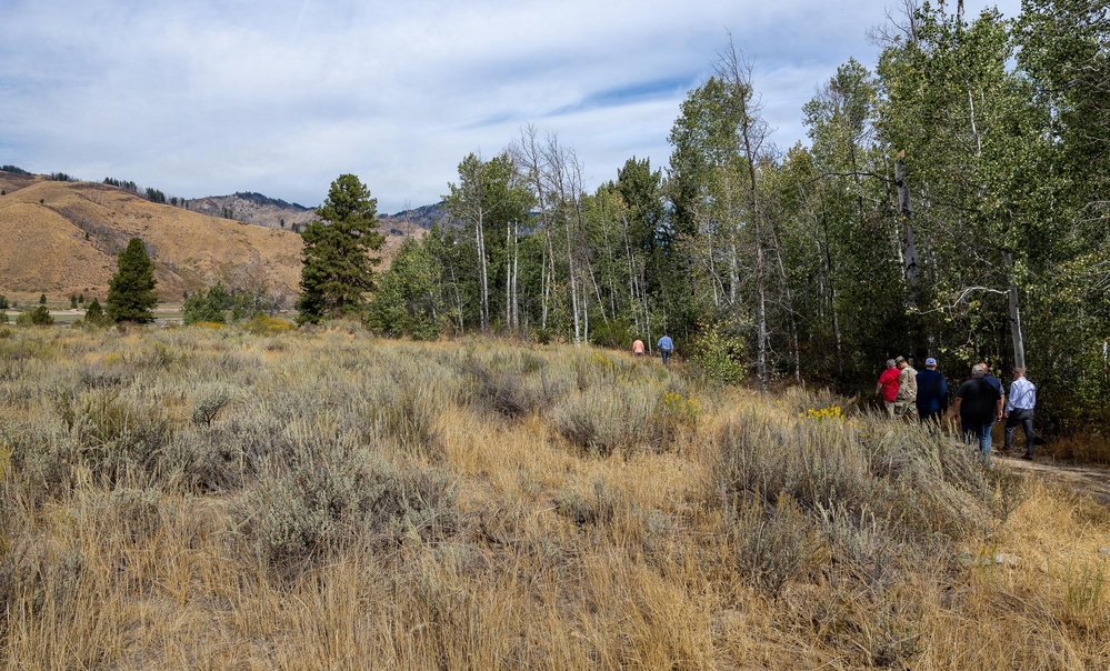 A group of people are walking along a dirt path through a high-elevation landscape characterized by sagebrush, dry grasses, and aspen trees. The terrain features golden-brown hillsides in the background, suggesting late summer or early autumn conditions. The scene appears to be a guided outdoor tour or field visit in a western mountain environment.