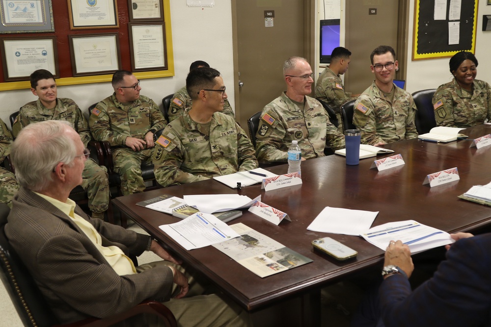 A group of U.S. Army soldiers in OCP (Operational Camouflage Pattern) uniforms are seated around a large conference table alongside at least one civilian, appearing to be engaged in a formal briefing or meeting. Name placards with small American flag decorations are visible on the table along with documents, notebooks, and personal items. The setting appears to be a military office or conference room with framed certificates and a bulletin board visible on the walls.