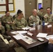 A group of U.S. Army soldiers in OCP (Operational Camouflage Pattern) uniforms are seated around a large conference table alongside at least one civilian, appearing to be engaged in a formal briefing or meeting. Name placards with small American flag decorations are visible on the table along with documents, notebooks, and personal items. The setting appears to be a military office or conference room with framed certificates and a bulletin board visible on the walls.