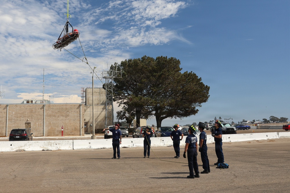A group of personnel in dark blue uniforms are conducting what appears to be a helicopter hoist rescue training exercise in an open paved area. A rescue basket (Stokes litter) with a person inside is suspended in the air from a cable, while ground crew members manage guide ropes and observe. The setting appears to be a military or emergency services facility based on the buildings, communications towers, and uniformed personnel visible.