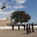 A group of personnel in dark blue uniforms are conducting what appears to be a helicopter hoist rescue training exercise in an open paved area. A rescue basket (Stokes litter) with a person inside is suspended in the air from a cable, while ground crew members manage guide ropes and observe. The setting appears to be a military or emergency services facility based on the buildings, communications towers, and uniformed personnel visible.