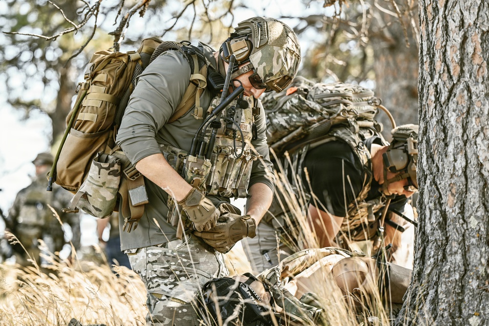 The image shows military or tactical personnel in a wooded outdoor environment, wearing full combat gear including helmets, plate carriers, and large rucksacks. The primary figure in the foreground appears to be attending to something or someone on the ground, possibly conducting a casualty care or equipment check scenario. A third individual is visible in the background, also in tactical attire.