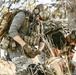 The image shows military or tactical personnel in a wooded outdoor environment, wearing full combat gear including helmets, plate carriers, and large rucksacks. The primary figure in the foreground appears to be attending to something or someone on the ground, possibly conducting a casualty care or equipment check scenario. A third individual is visible in the background, also in tactical attire.