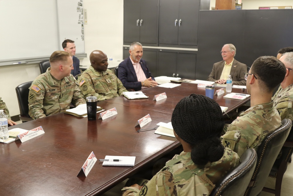 A formal meeting or briefing is taking place around a large conference table, involving several U.S. Army soldiers in OCP (Operational Camouflage Pattern) uniforms and civilian attendees in business attire. Name placards with American flag designs are visible on the table, suggesting an official or structured engagement. The setting appears to be a military installation conference room.