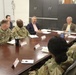 A formal meeting or briefing is taking place around a large conference table, involving several U.S. Army soldiers in OCP (Operational Camouflage Pattern) uniforms and civilian attendees in business attire. Name placards with American flag designs are visible on the table, suggesting an official or structured engagement. The setting appears to be a military installation conference room.