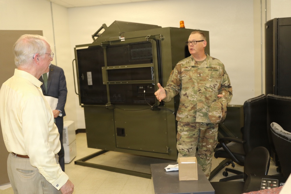 A U.S. Army soldier in MultiCam/OCP camouflage uniform is briefing or presenting to two civilians in an indoor facility. The soldier appears to be explaining a large olive-drab military equipment unit, possibly a ruggedized electronics or communications shelter. The setting appears to be a demonstration or tour of military equipment.